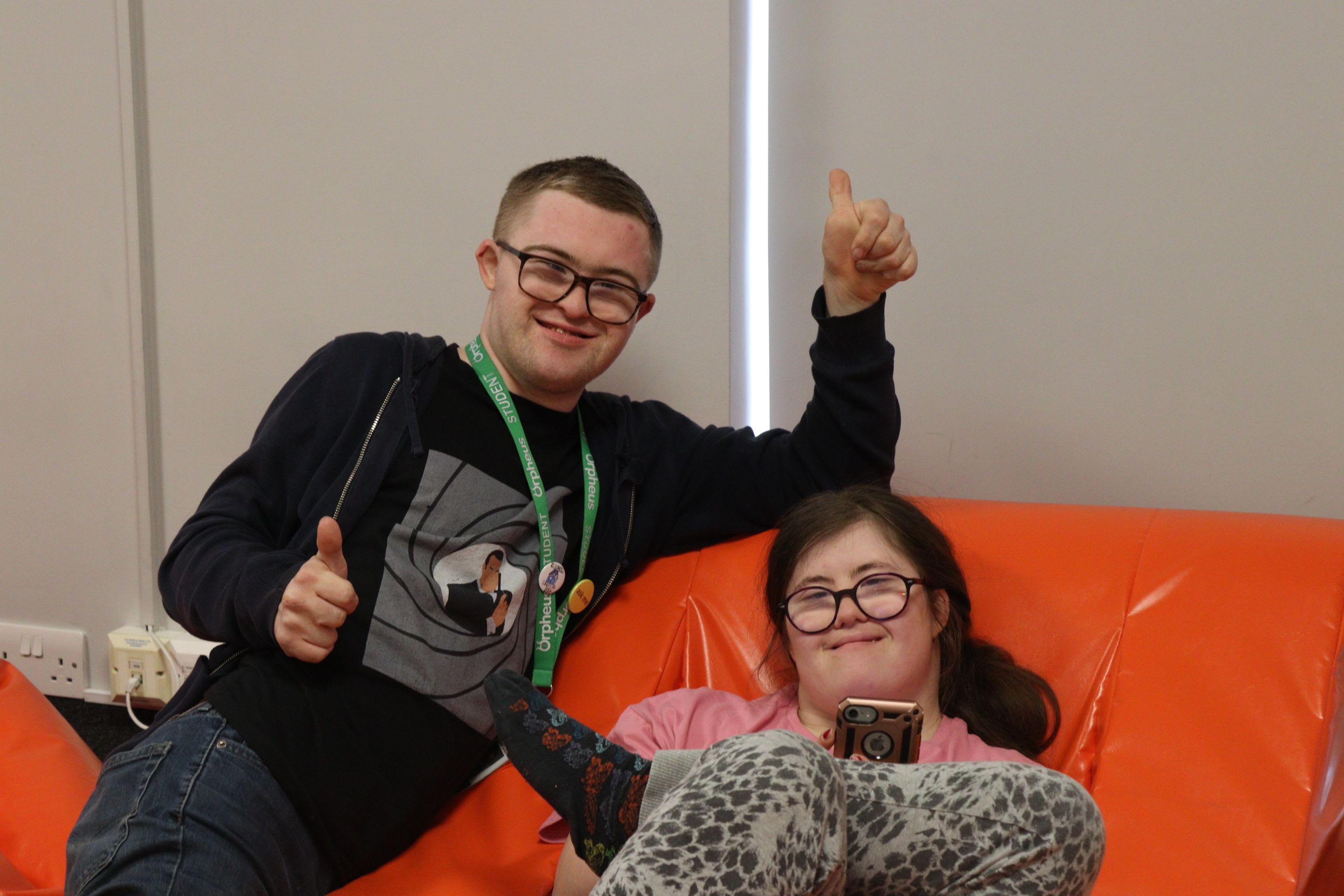 Two students sat on a low orange chair in the sensory room at the Orpheus centre, in Godstone, Surrey. They are both smiling and one of the students is giving the thumbs up sign.