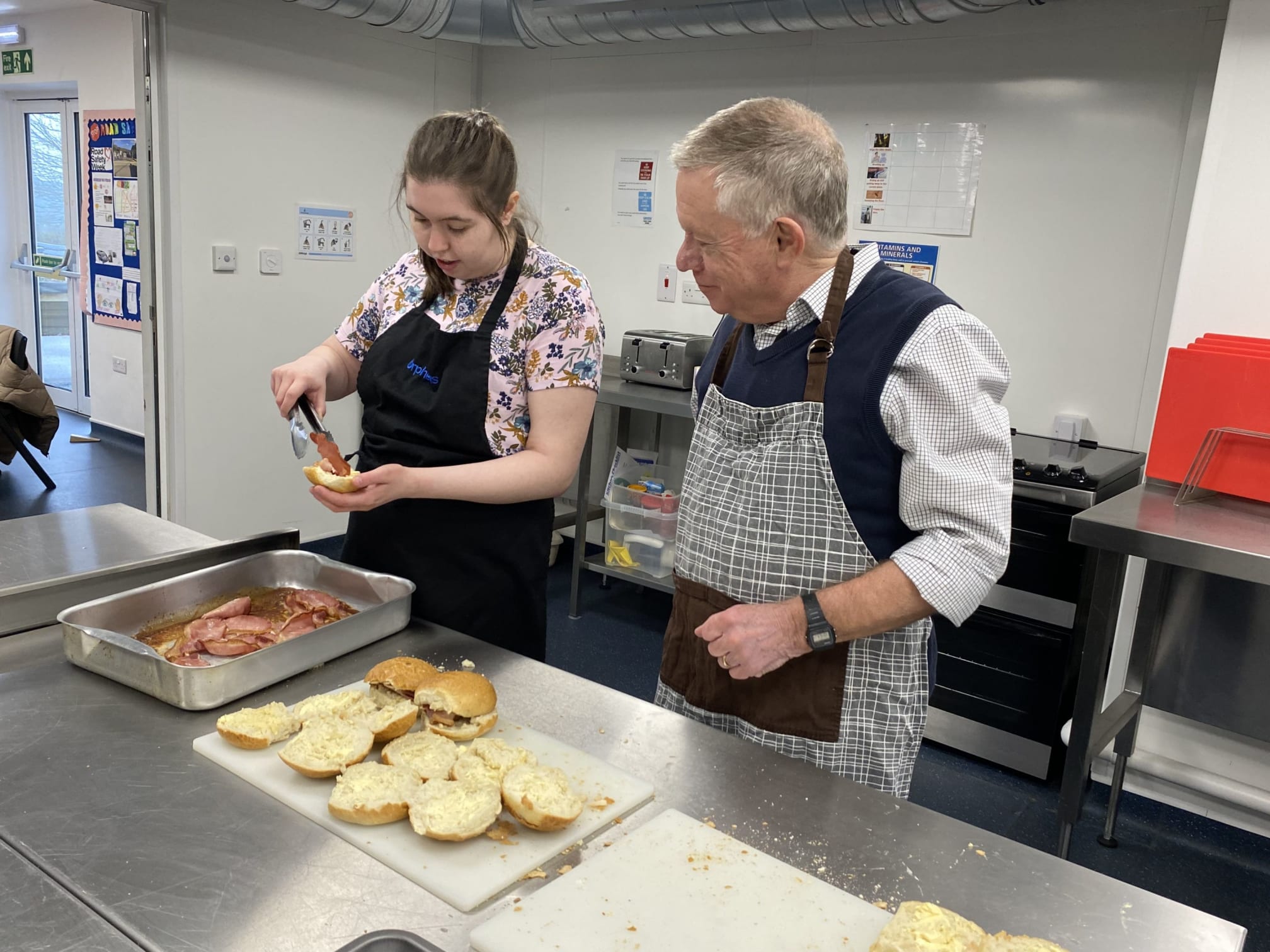 Orpheus student Lauren and volunteer Steve, both standing at a counter in the life skills kitchen, Steve assisting Lauren who is adding bacon to bread rolls
